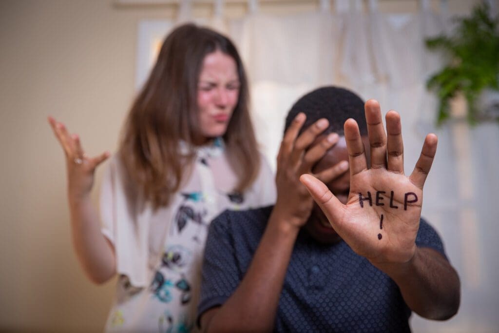 A person sits with their head in their hand, showing "HELP!" written on their palm, while another stands behind them appearing frustrated—capturing the urgent need for support that many nonprofit organizations address. AlternativesBeauty.org