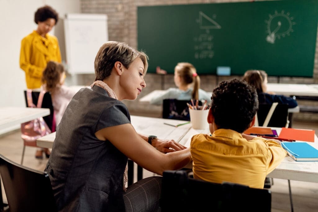 A teacher helps a student at a desk in a classroom at a nonprofit, while another teacher and students are near a chalkboard with math diagrams in the background. AlternativesBeauty.org
