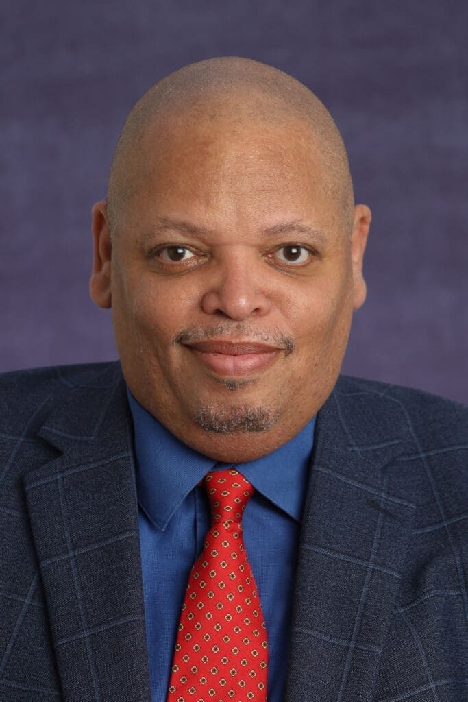 A man in a blue suit jacket, blue shirt, and red patterned tie poses for a formal portrait against a purple background, reflecting the professionalism often seen in nonprofit leadership. AlternativesBeauty.org