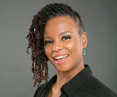 Woman with dark, curly hair styled to one side, wearing a black top and blue earring, smiling in front of a plain gray background—her confident presence reflects her inspiring work in the nonprofit sector. AlternativesBeauty.org